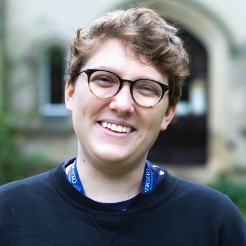 Hal smiling at the camera. He has brown curly hair, glasses, and wears a black jumper.