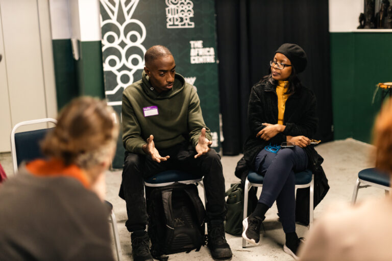 People sitting around in a circle. In the middle is a Black man with short hair and a green jumper speaking to the group. In the background is a banner for the Africa Centre, where the event is hosted.