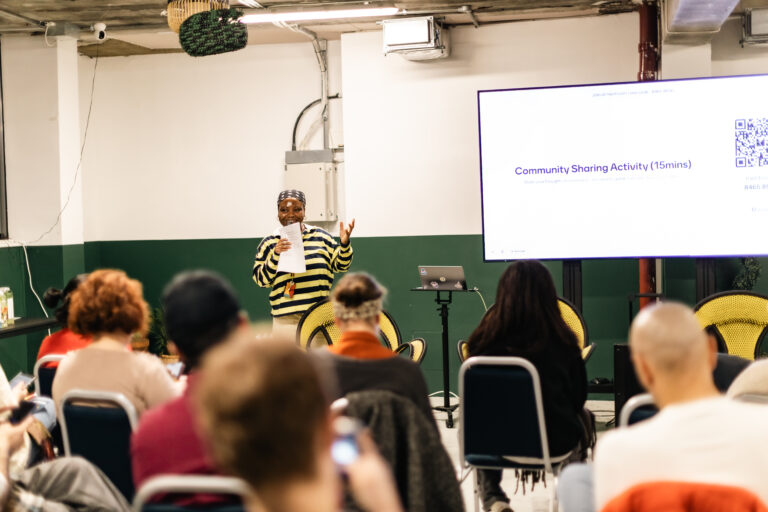 Rows of people facing towards a OUTpatients Volunteer Lead, Jawadat, at the front of the room. Jawadat, OUTpatients' Volunteer Coordinator, is smiling and speaking into a microphone. Next to her is a powerpoint slide that reads ‘Community Sharing Activity (15 mins)’. She is a Black woman wearing a black and white striped top, hoop earrings and a head scarf.
