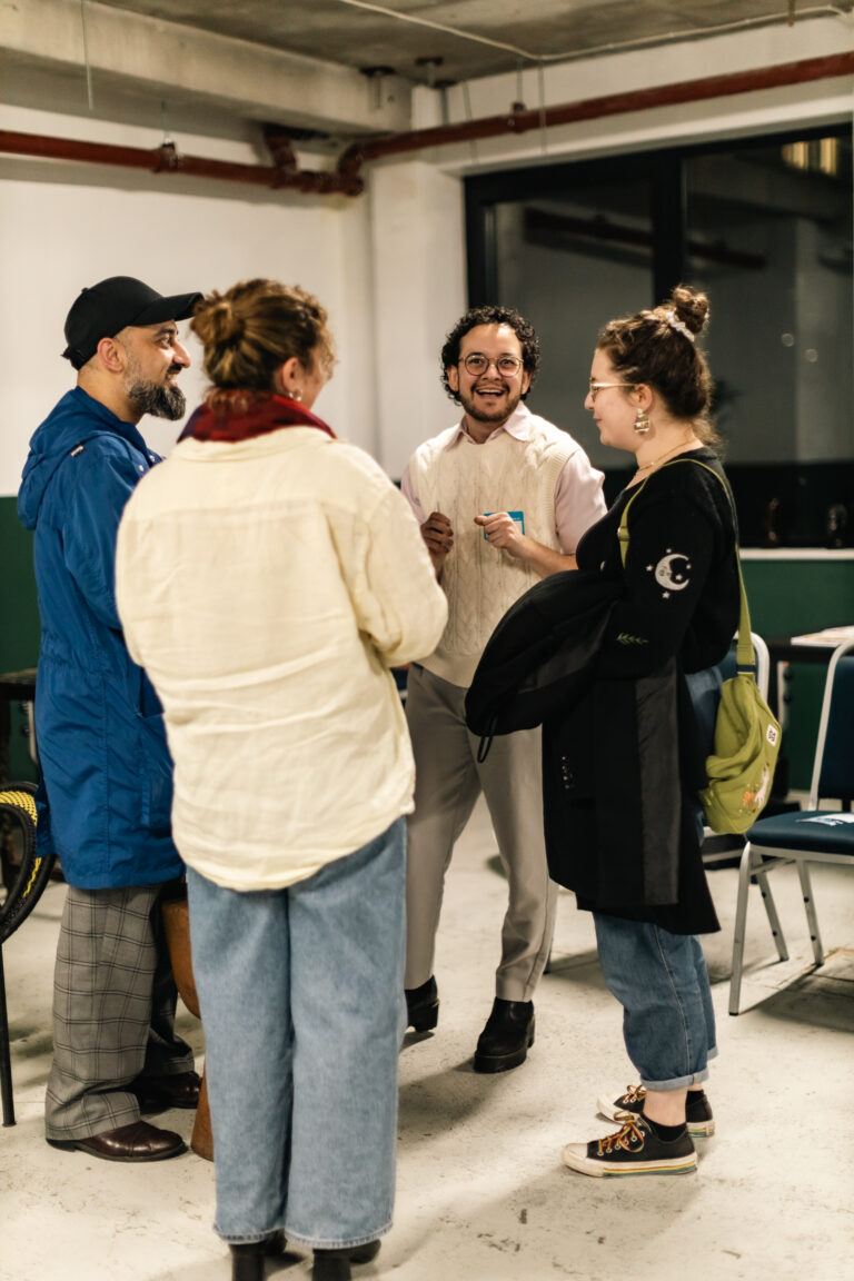 Photo of 4 people standing in a circle, smiling and talking. On the left is a South Asian man with a beard, a long blue coat, and a black cap. On the right is a Jewish man with curly hair, a beard, and a white sweater vest.
