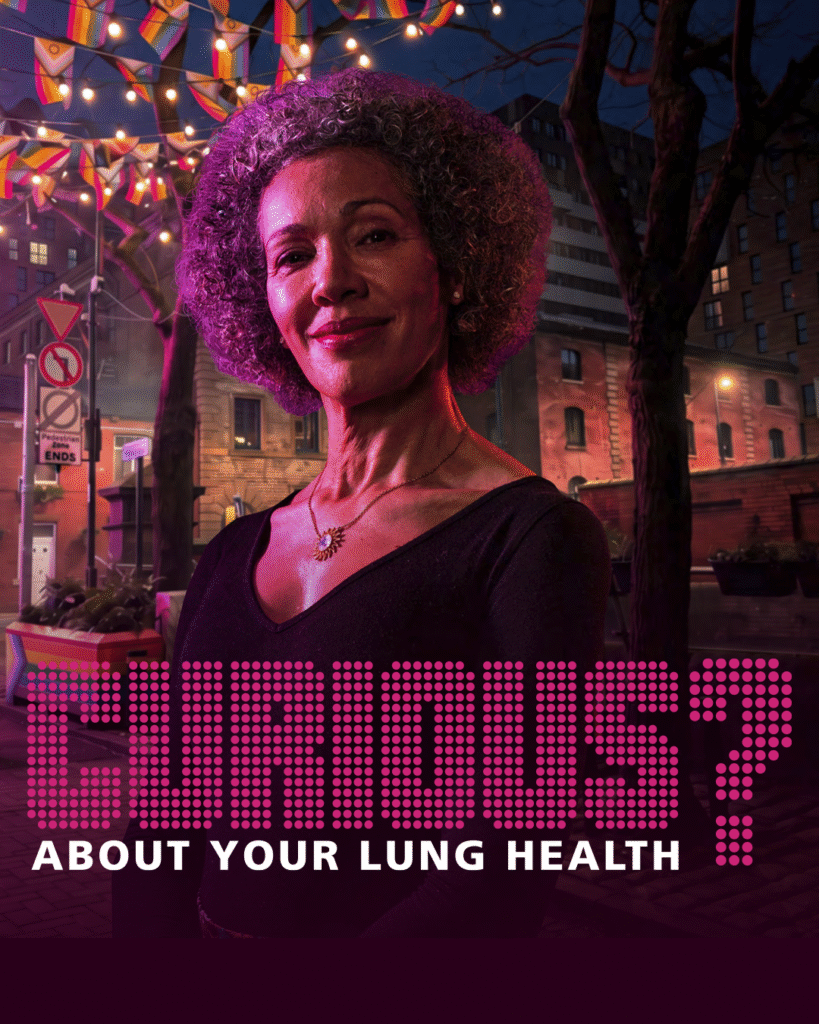 Curious about your lung health? A Black woman smiles into the camera. In the background is Canal Street in Manchester, with Pride flag bunting handing in the trees.