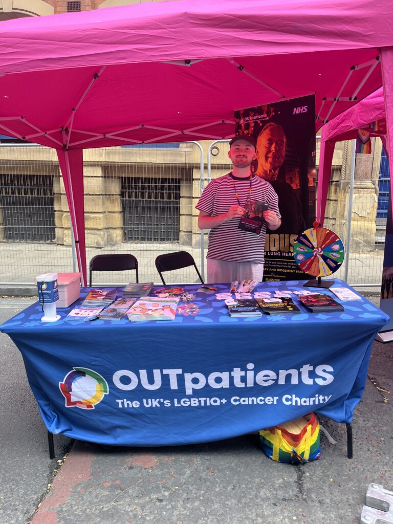 Our Comms and Public Affairs Manager, Ezra, standing behind the OUTpatients Pride stall. The table is covered in booklets and resources.