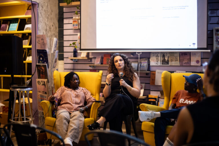 Professor Sheeren Nabhani-Gebara holds a microphone and speaks to a room of people alongside other panelists. They are sitting in comfy looking yellow armchairs in Common Press bookshop.